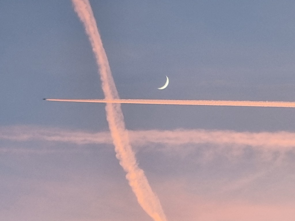 Un cer violet cu lună în formă de semilună și două urme de siaj de la avioane: o urmă orizontală, sub semiluna albă și o urmă verticală, ușor înclinată și mai cețoasă, deja mai împrăștiată.