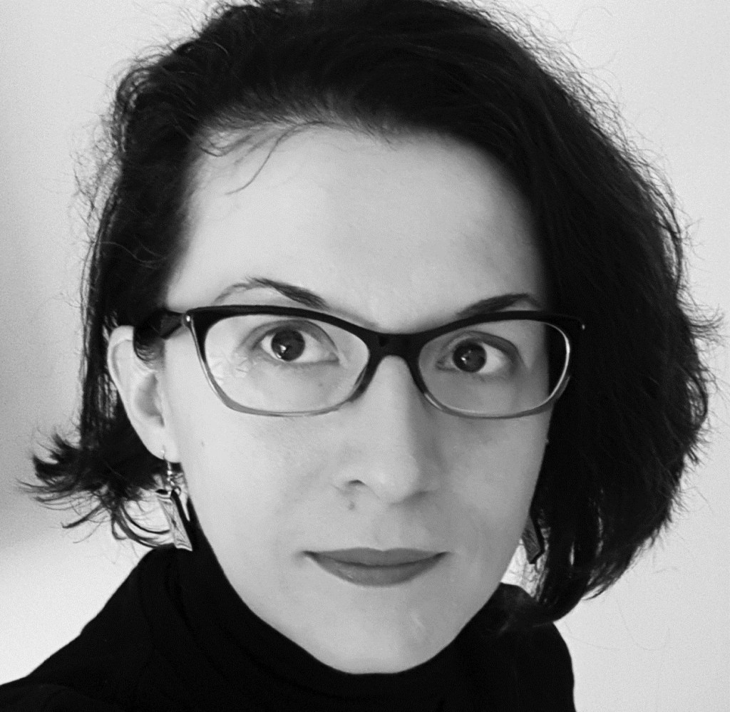 Portret alb-negru al arhitectei Oana Mihăescu, persoană brunetă, cu părul mediu, tuns asimetric, cu ochelari și privire curioasă și interesată.